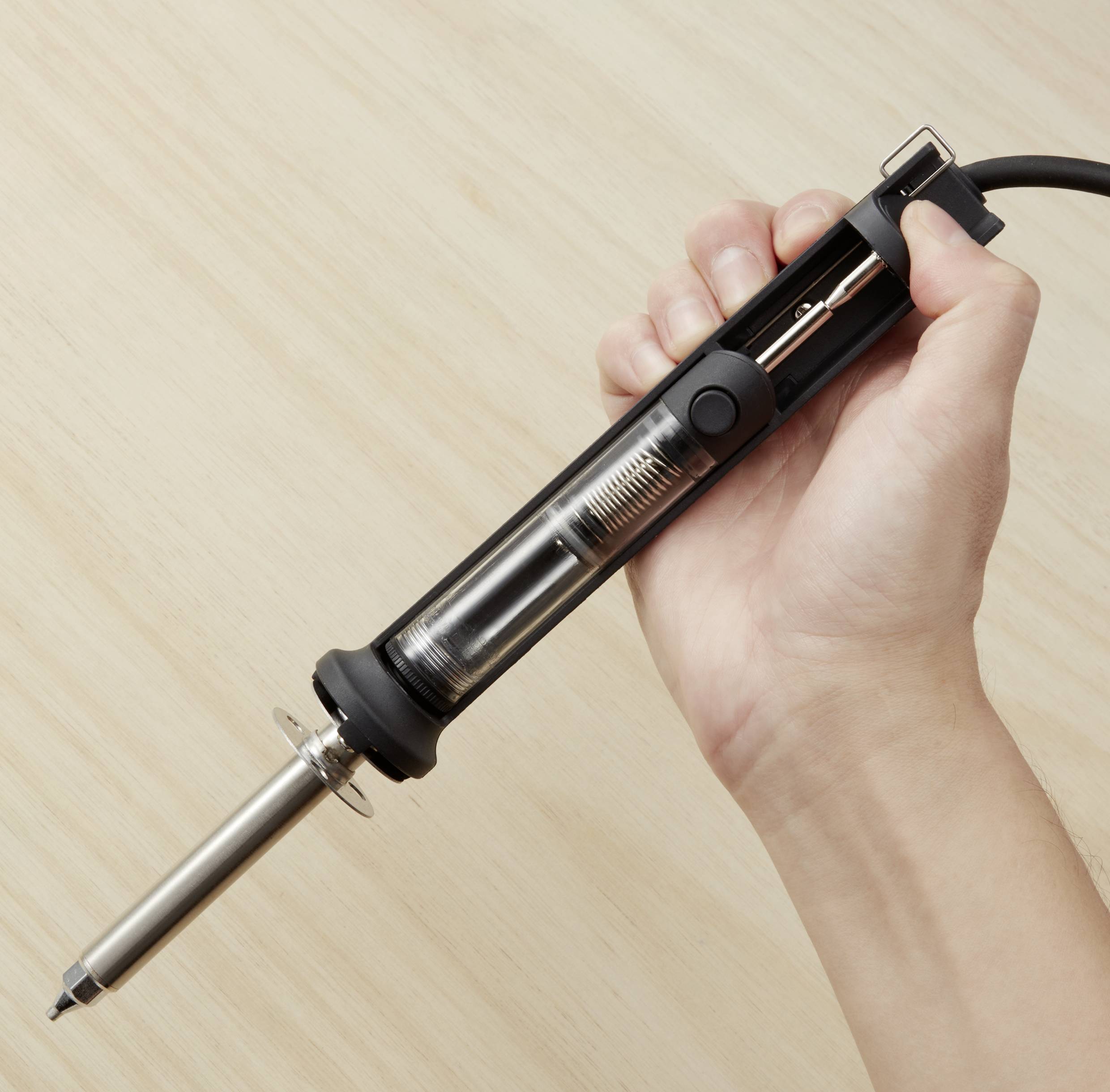 Une main tient un fer à souder au-dessus d'une table en bois. Le fer à souder a une poignée noire et une pointe en métal.