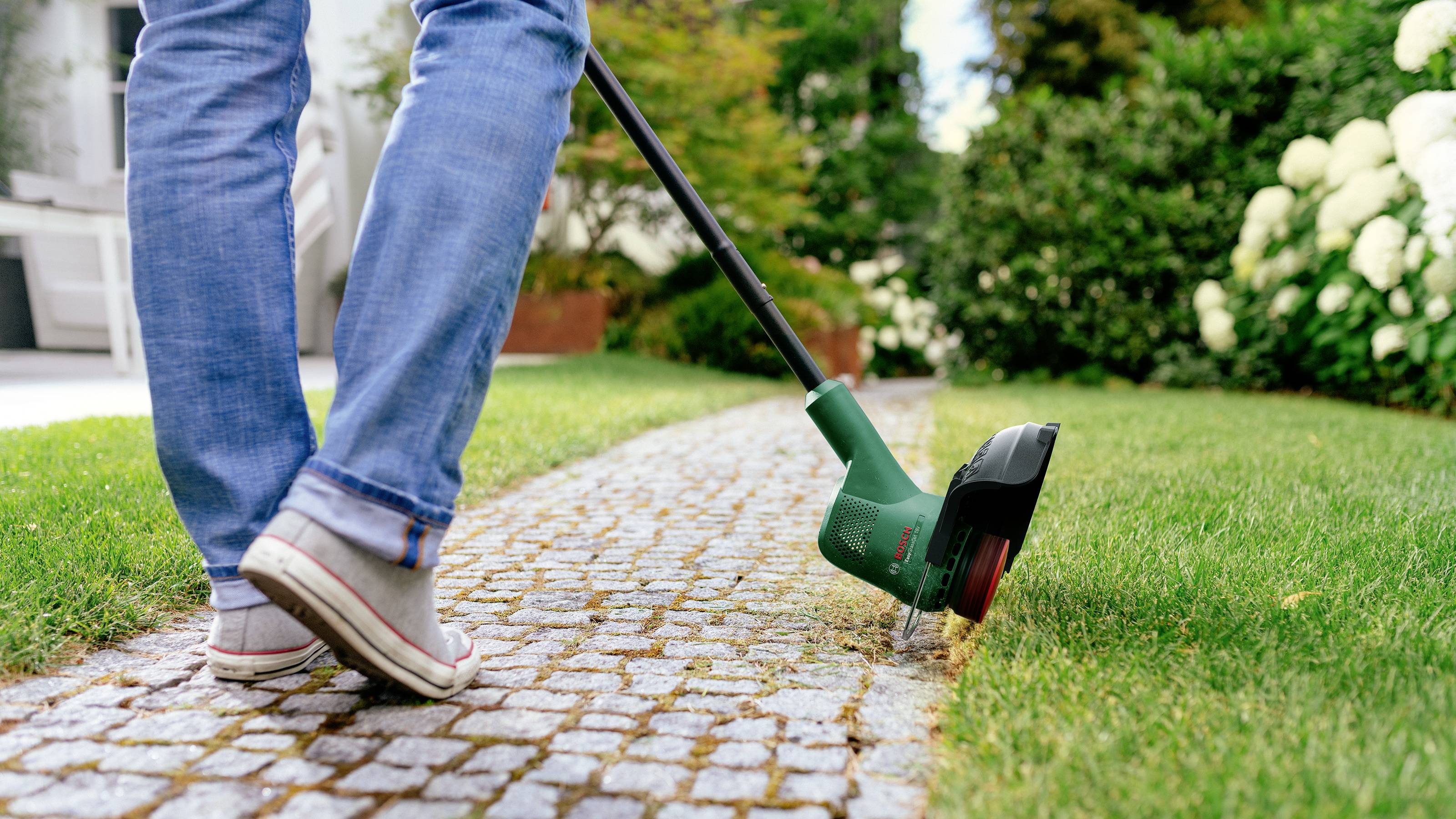 Une personne portant un jean et des baskets taille le bord d'une pelouse bien entretenue avec un coupe-bordure électrique le long d'un chemin pavé.