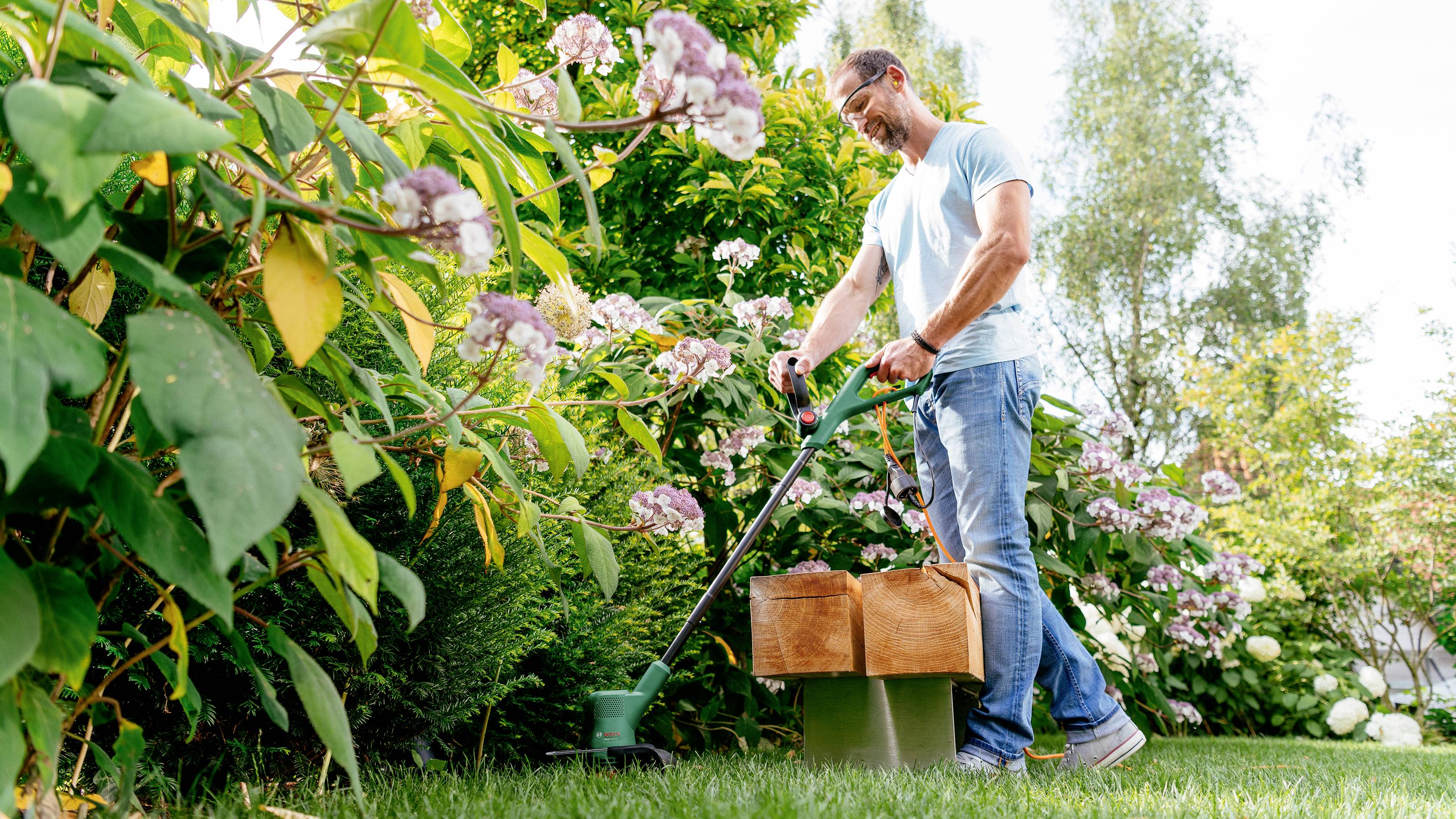 Un homme tond la pelouse dans un jardin en fleurs. Il porte des lunettes et des vêtements décontractés.