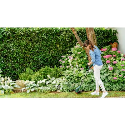 Une femme en tenue décontractée utilise un coupe-bordures dans le jardin. À l'arrière-plan, on aperçoit des plantes vertes et des hortensias roses.