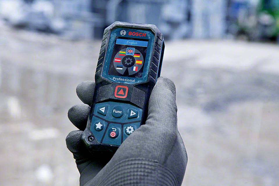 Une main tient un télémètre laser Bosch avec affichage et commandes. Arrière-plan flou, scène de travail.