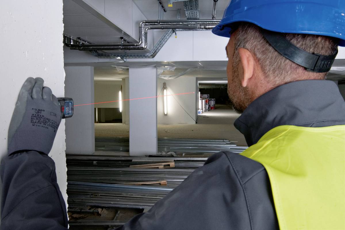 Un ouvrier portant un casque et un gilet de sécurité mesure la distance à l'intérieur d'un bâtiment à l'aide d'un télémètre laser.