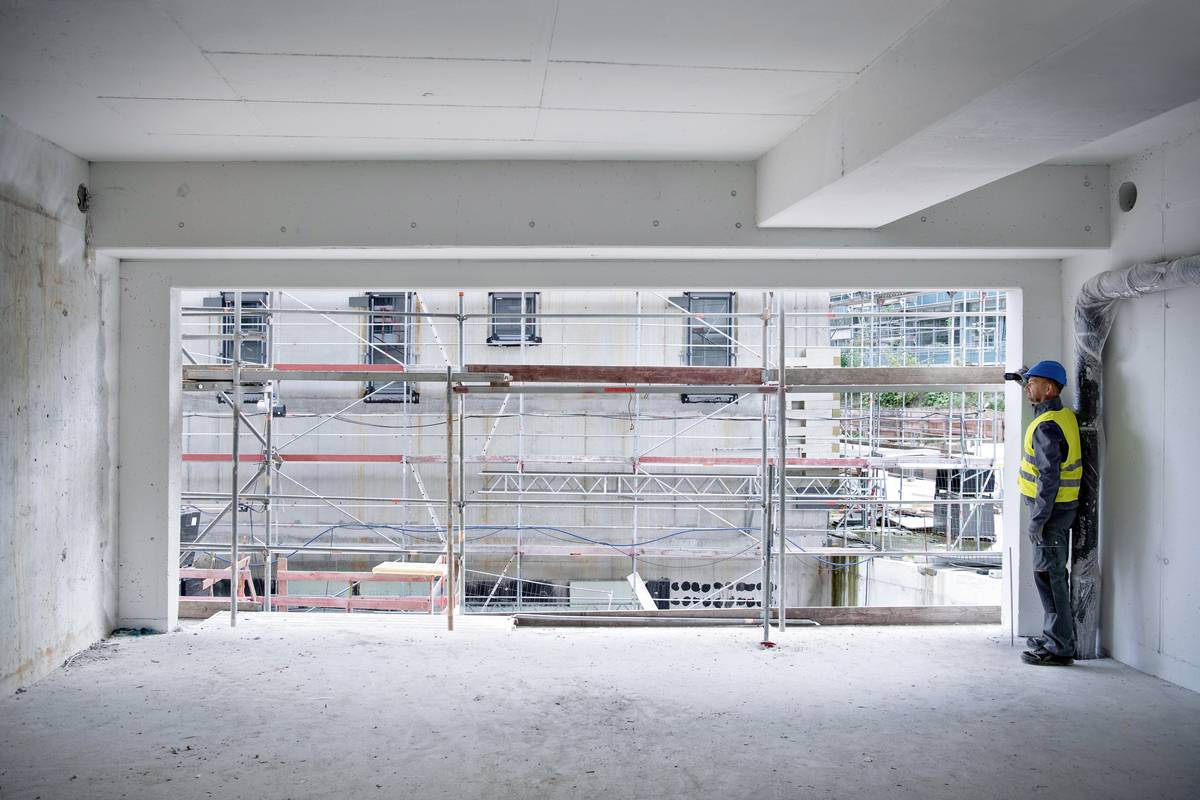 Un ouvrier portant un casque se tient dans une pièce inachevée à côté d'un grand cadre de fenêtre. À l'extérieur, un échafaudage est visible.