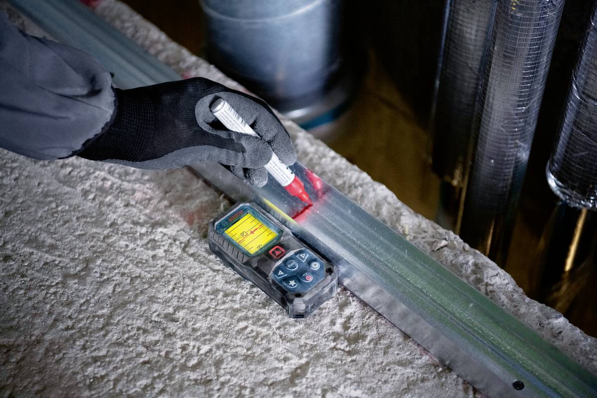 Une personne portant des gants utilisant un appareil de mesure numérique pour mesurer la longueur d'une poutre métallique dans un environnement industriel.