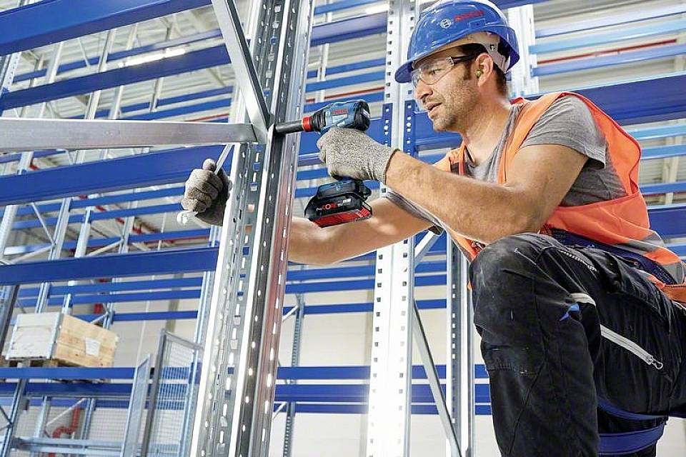 Un ouvrier sur un chantier fixe des poutrelles métalliques à l'aide d'une visseuse sans fil. Il porte un équipement de sécurité, notamment un casque et un gilet de protection.