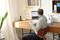 Un homme est assis à un bureau et travaille sur un ordinateur portable. La pièce est bien éclairée, avec des plantes et du matériel de bureau autour de lui.