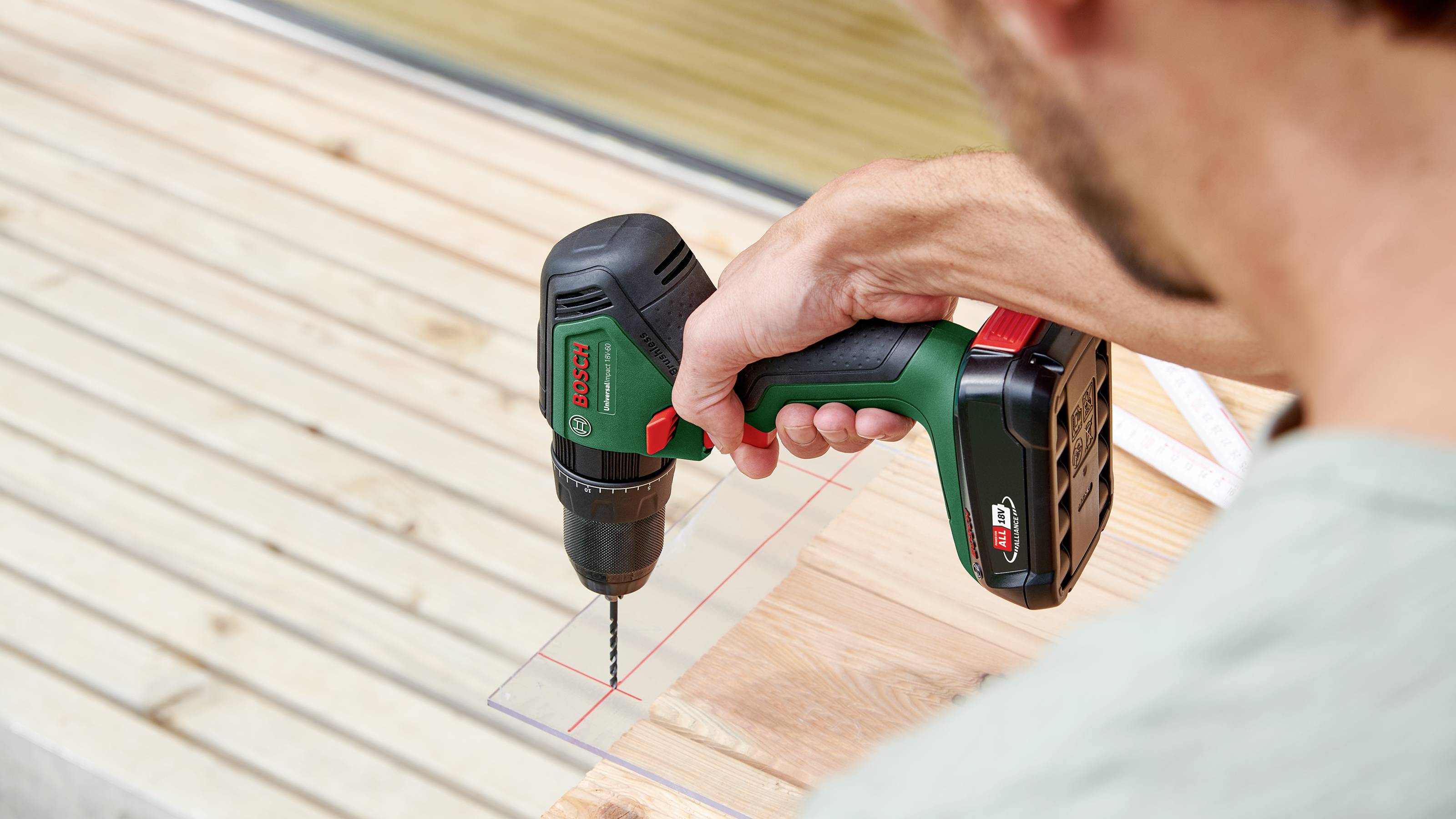 Une personne utilise une perceuse électrique verte et noire pour travailler une planche de bois. Un sol en bois est visible en arrière-plan.