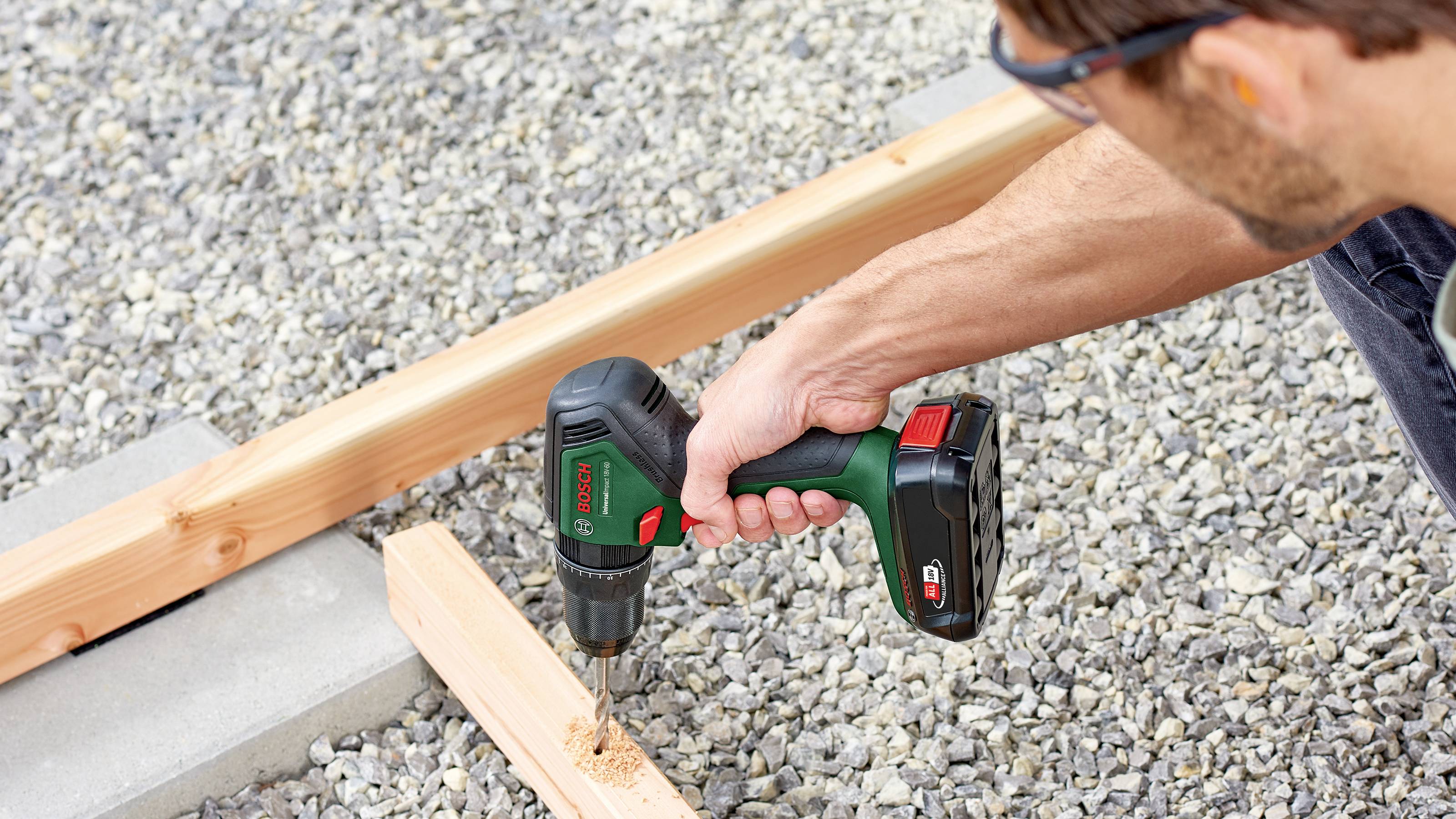 Une personne fore une planche en bois avec une visseuse-perceuse sans fil sur un lit de gravier. Des copeaux de bois sont produits.