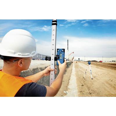 Un ouvrier portant un casque et un gilet de sécurité effectue un relevé avec un niveau de chantier sur un site de construction. Des travaux de construction sont visibles en arrière-plan.