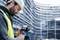 Un ouvrier portant un casque de sécurité et un gilet de signalisation utilise un appareil de mesure laser sur un chantier devant un bâtiment échafaudé.