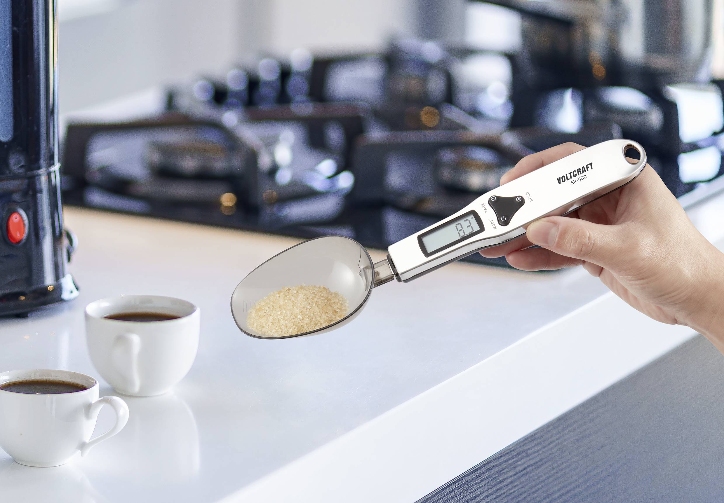 Une personne tient une cuillère numérique avec un écran affichant un poids. Deux tasses à café sont posées en arrière-plan sur un plan de travail de cuisine.