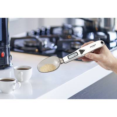 Une personne tient une cuillère numérique avec un écran affichant un poids. Deux tasses à café sont posées en arrière-plan sur un plan de travail de cuisine.