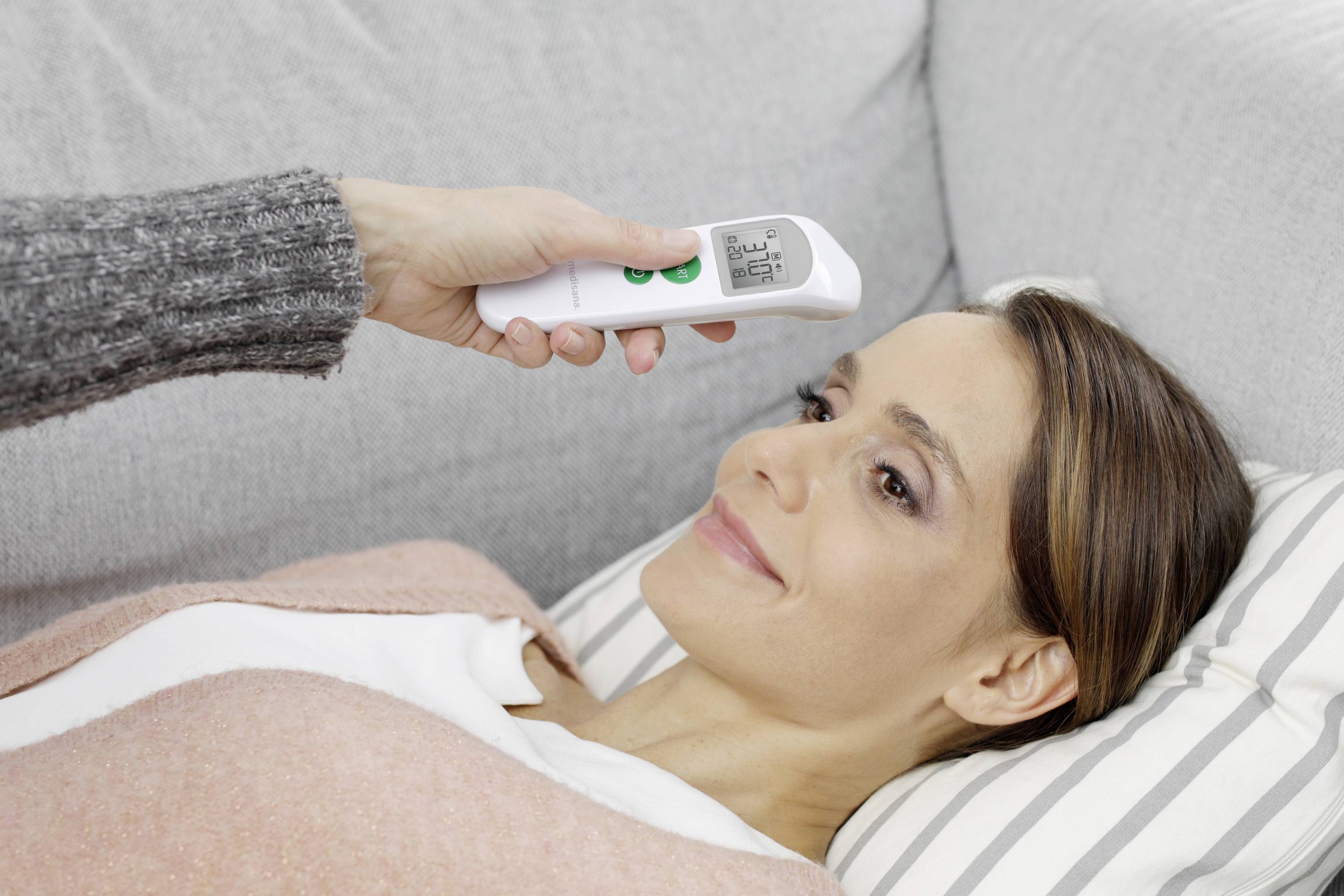 Une personne mesure la température d'une femme souriante à l'aide d'un thermomètre frontal alors qu'elle est allongée sur un canapé.