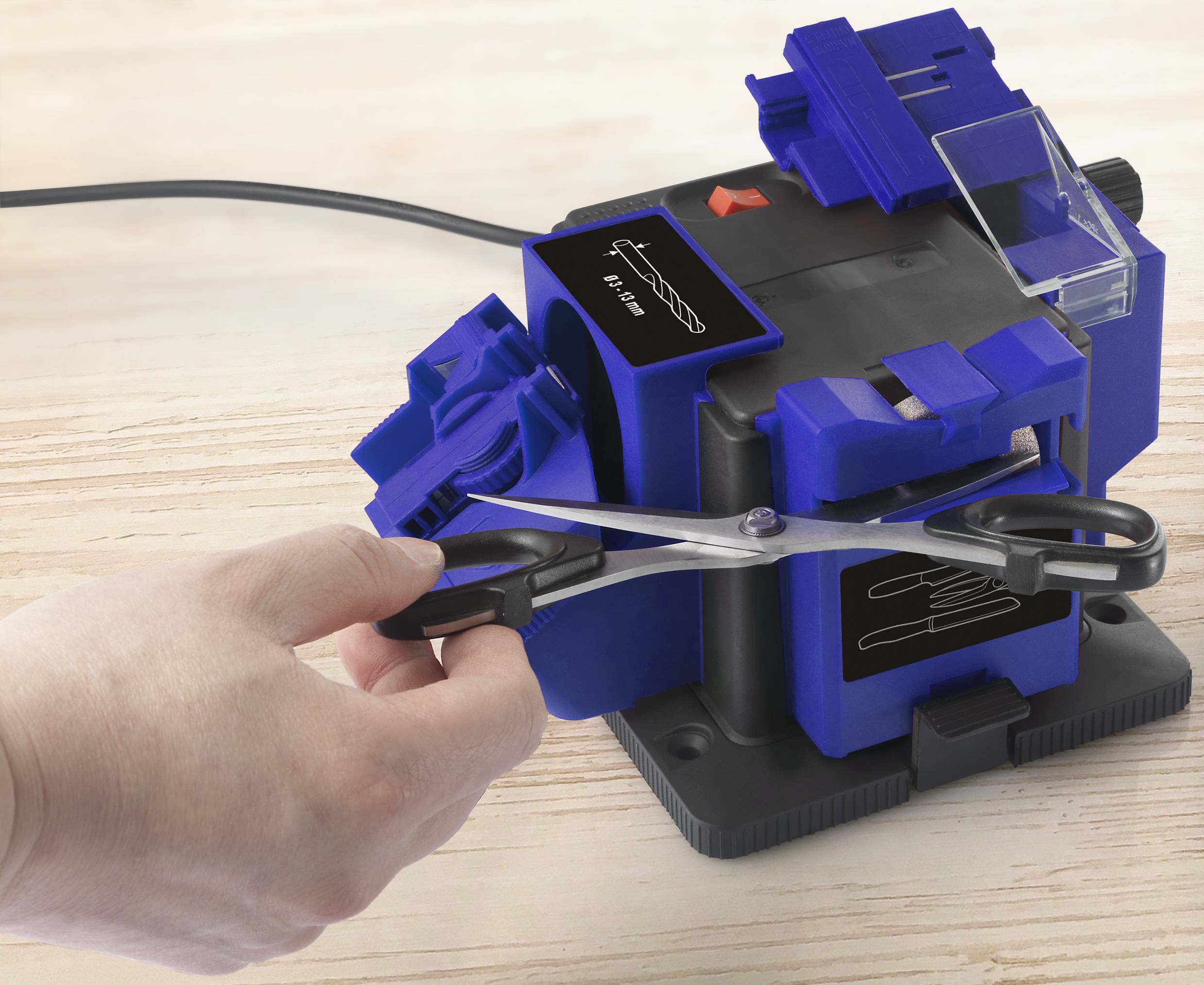 Une main aiguise des ciseaux avec un dispositif d'affûtage bleu et noir. L'appareil est posé sur une surface en bois.