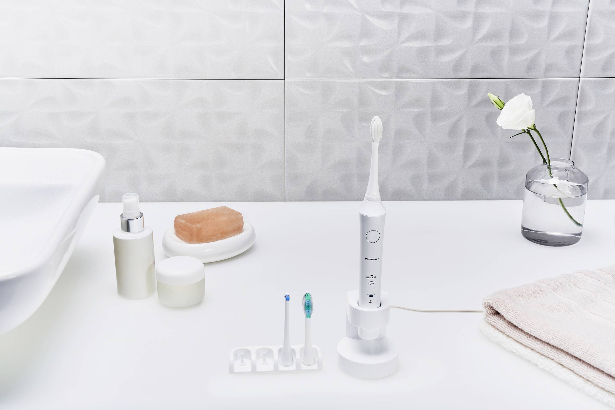 Brosse à dents électrique sur un lavabo blanc de salle de bain, accompagnée de têtes de brosse supplémentaires, de savon, d'une serviette et d'une fleur blanche dans un vase.