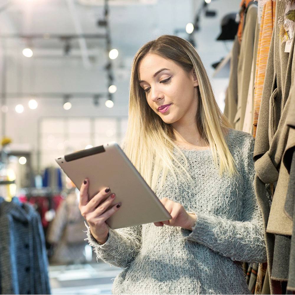 Une femme dans un magasin de vêtements regarde une tablette, entourée de vêtements suspendus à des portants. Elle semble concentrée.