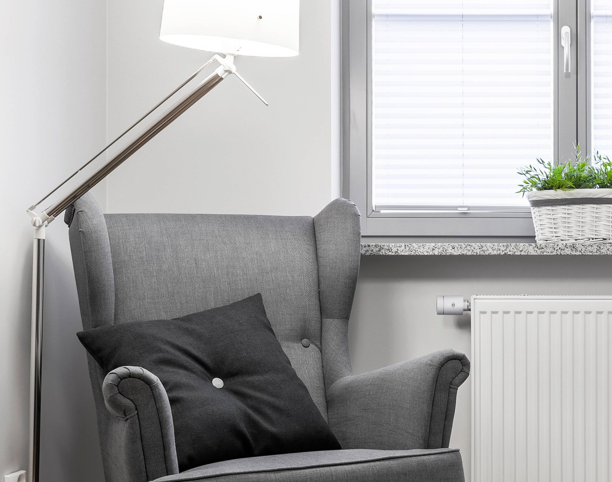 Fauteuil gris à oreilles avec coussin noir à côté d'un lampadaire ; près d'une fenêtre avec des stores blancs ; radiateur et plantes décoratives.