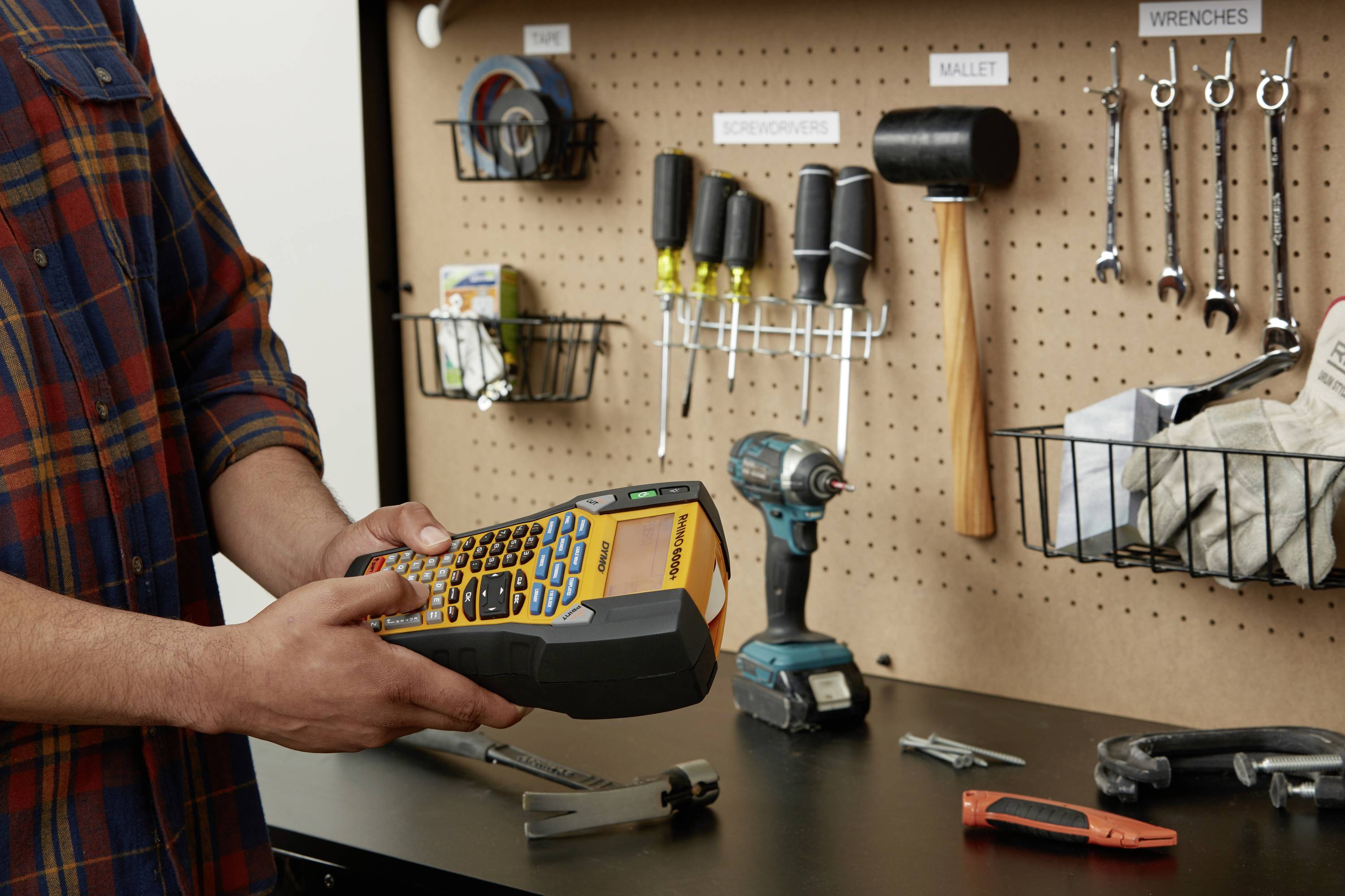 Un homme tient un appareil d'étiquetage dans un atelier. À l'arrière-plan, des outils tels que des perceuses, un marteau et des clés sont fixés au mur.