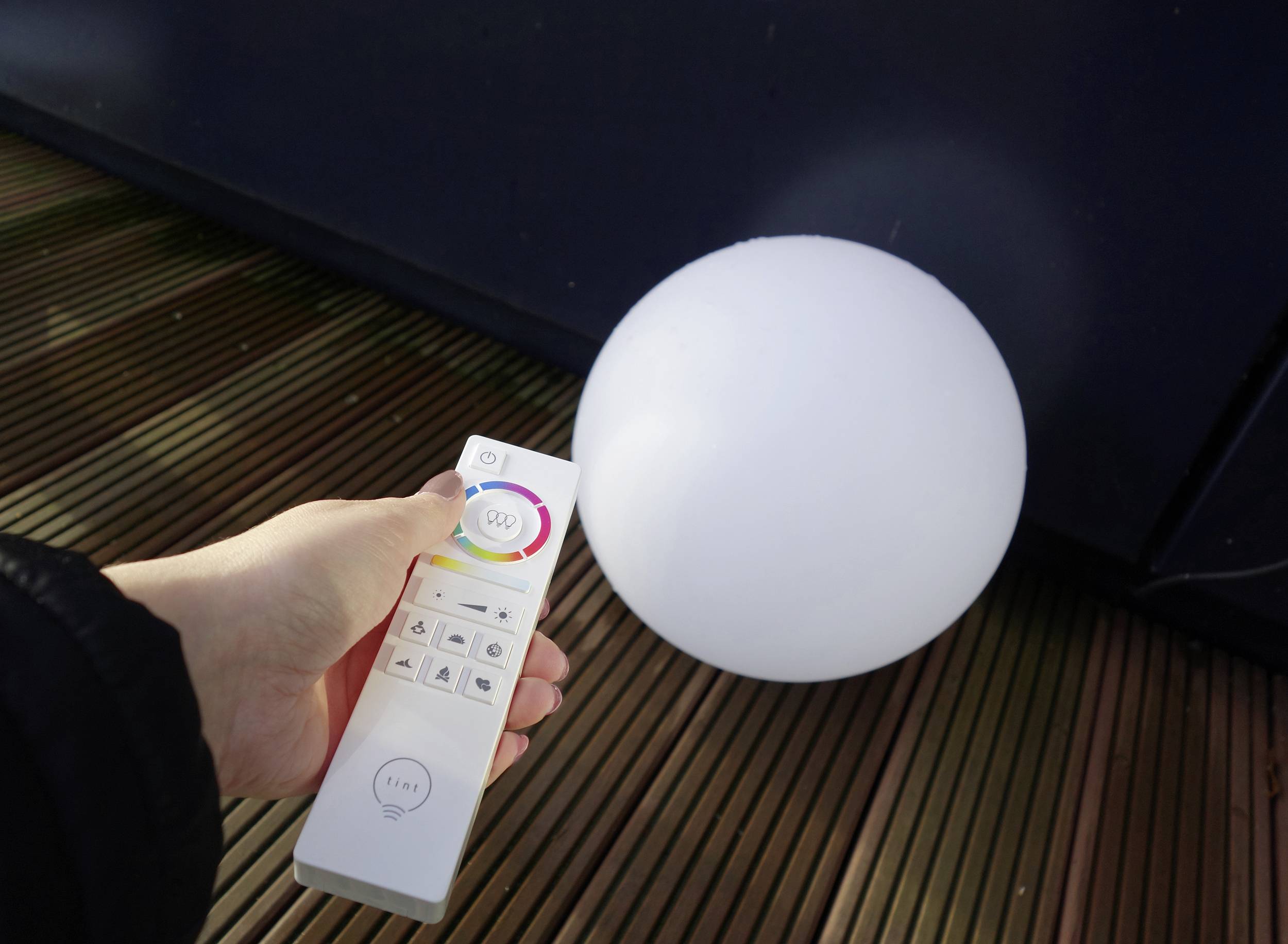 Une main tient une télécommande avec une roue de couleurs et des touches, la dirigeant vers un luminaire blanc et rond posé sur un sol en bois.