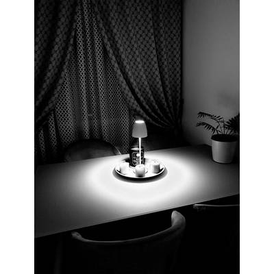 Une table avec une petite lampe éclairant un récipient à boisson et des tasses. Un rideau et une plante sont visibles en arrière-plan.
