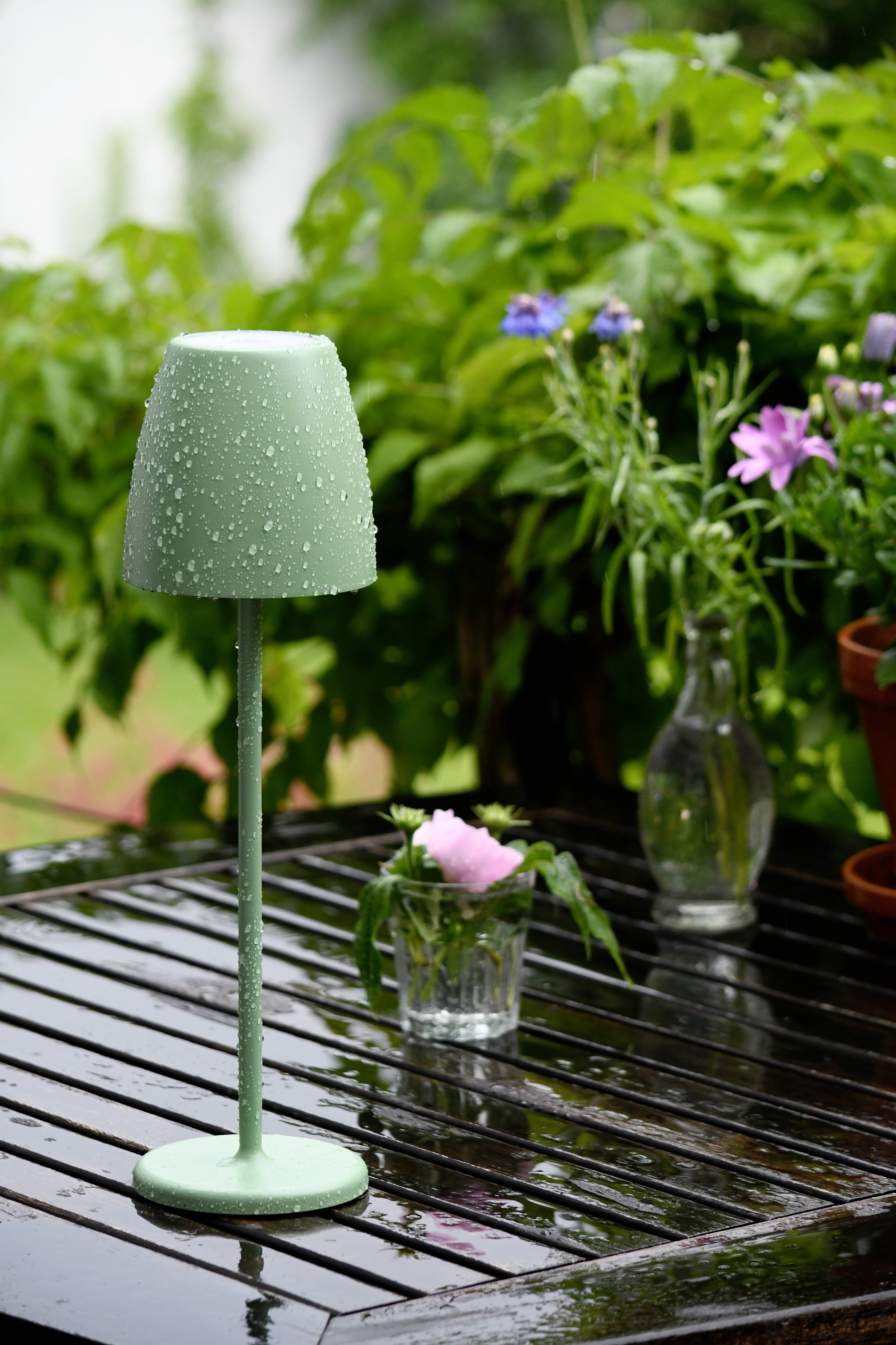 Une lampe humide et verte est posée sur une table mouillée par la pluie dans le jardin. En arrière-plan, on peut voir des plantes vertes et des fleurs dans des verres.