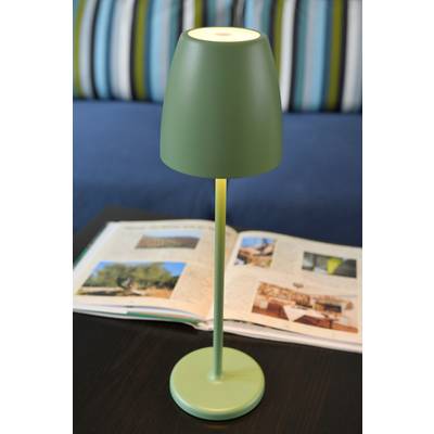 Lampe verte allumée sur une table. À l'arrière-plan, un livre ouvert avec des images est posé, et dans la pièce se trouve un canapé avec des coussins rayés.