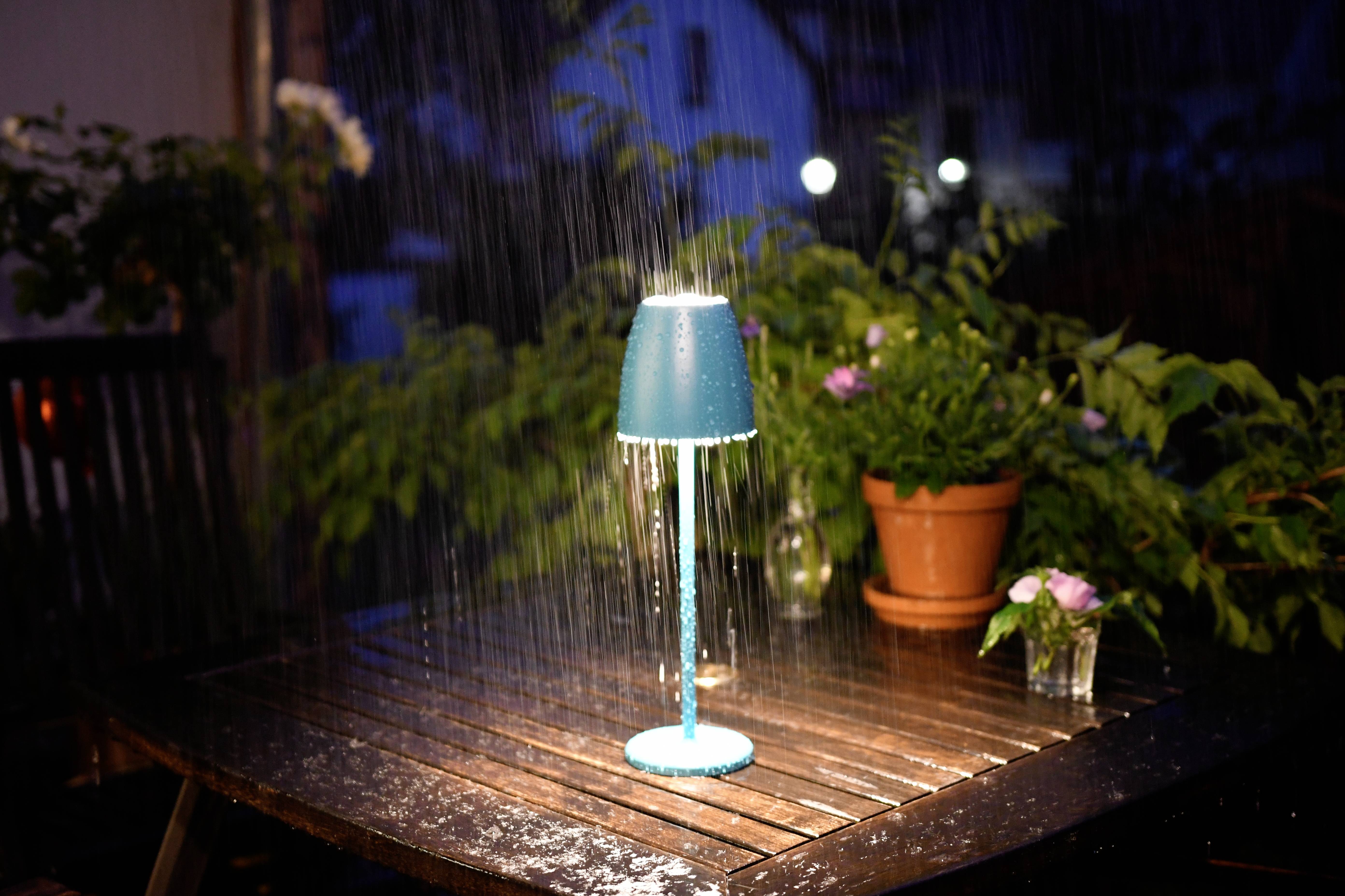 Une lampe est posée sur une table à l'extérieur et est arrosée par la pluie. Des plantes et des vases à fleurs sont également sur la table.