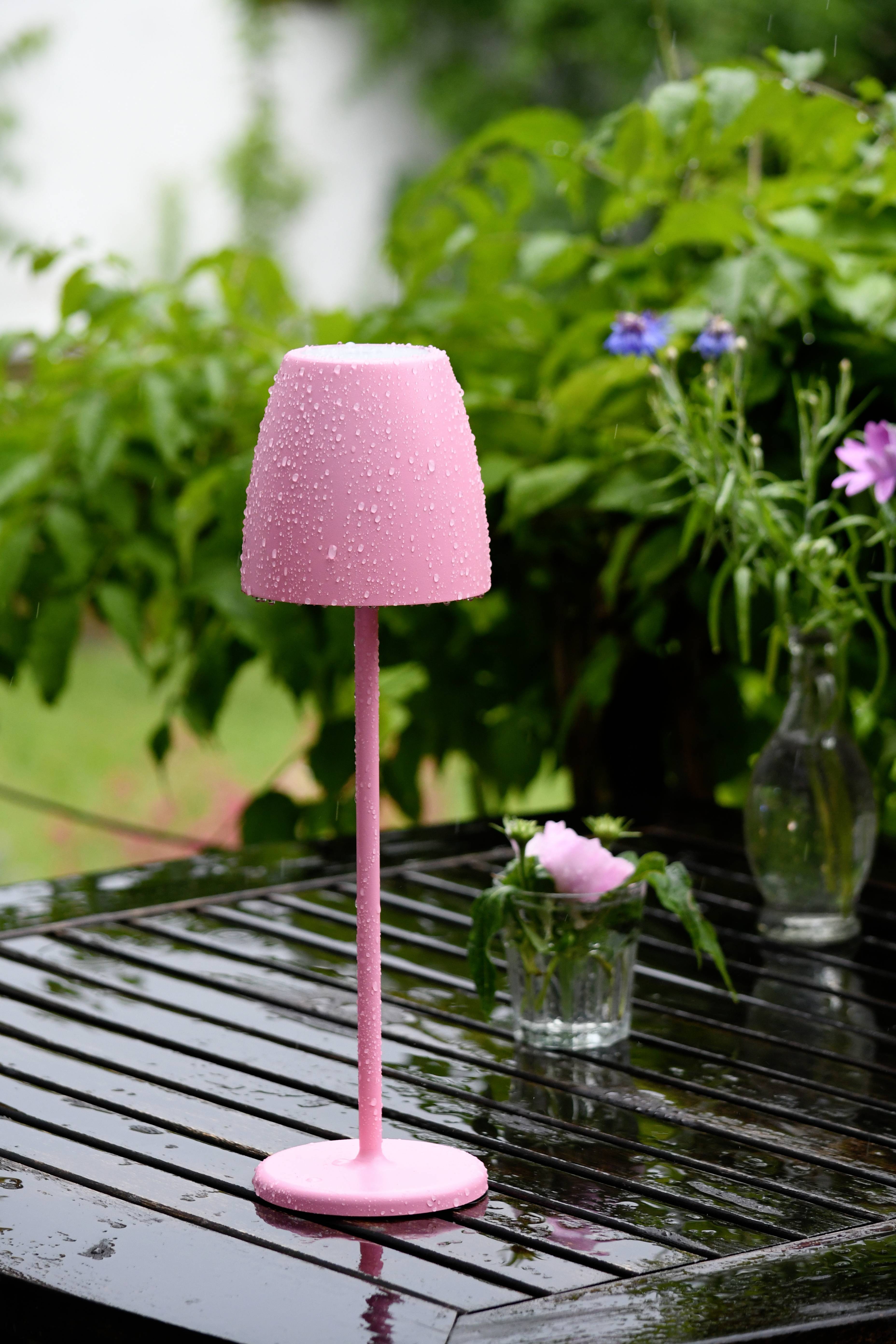 Une lampe rose avec des gouttes d'eau est posée sur une table en bois humide à l'extérieur. En arrière-plan, on aperçoit des plantes vertes et des fleurs.