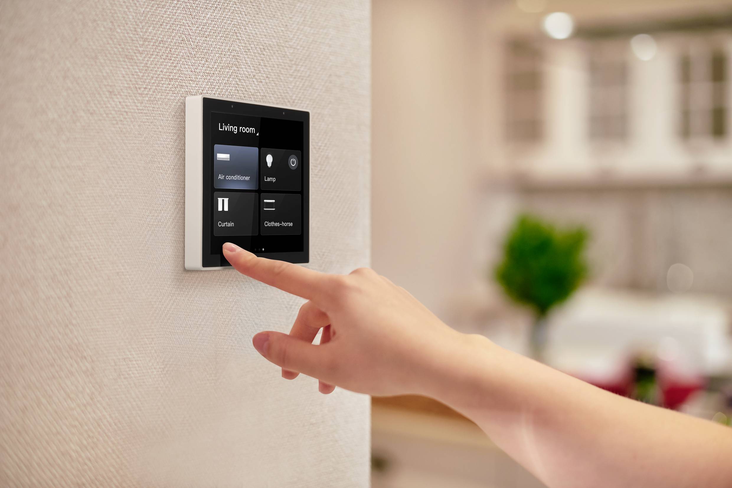 Une main actionne un écran domotique digital mural pour contrôler les appareils du salon, tels que la climatisation et les rideaux.