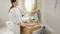 Une femme dans la salle de bain accroche une serviette à un crochet noir. La pièce est aménagée de manière moderne, avec un miroir rond et des carreaux verts.