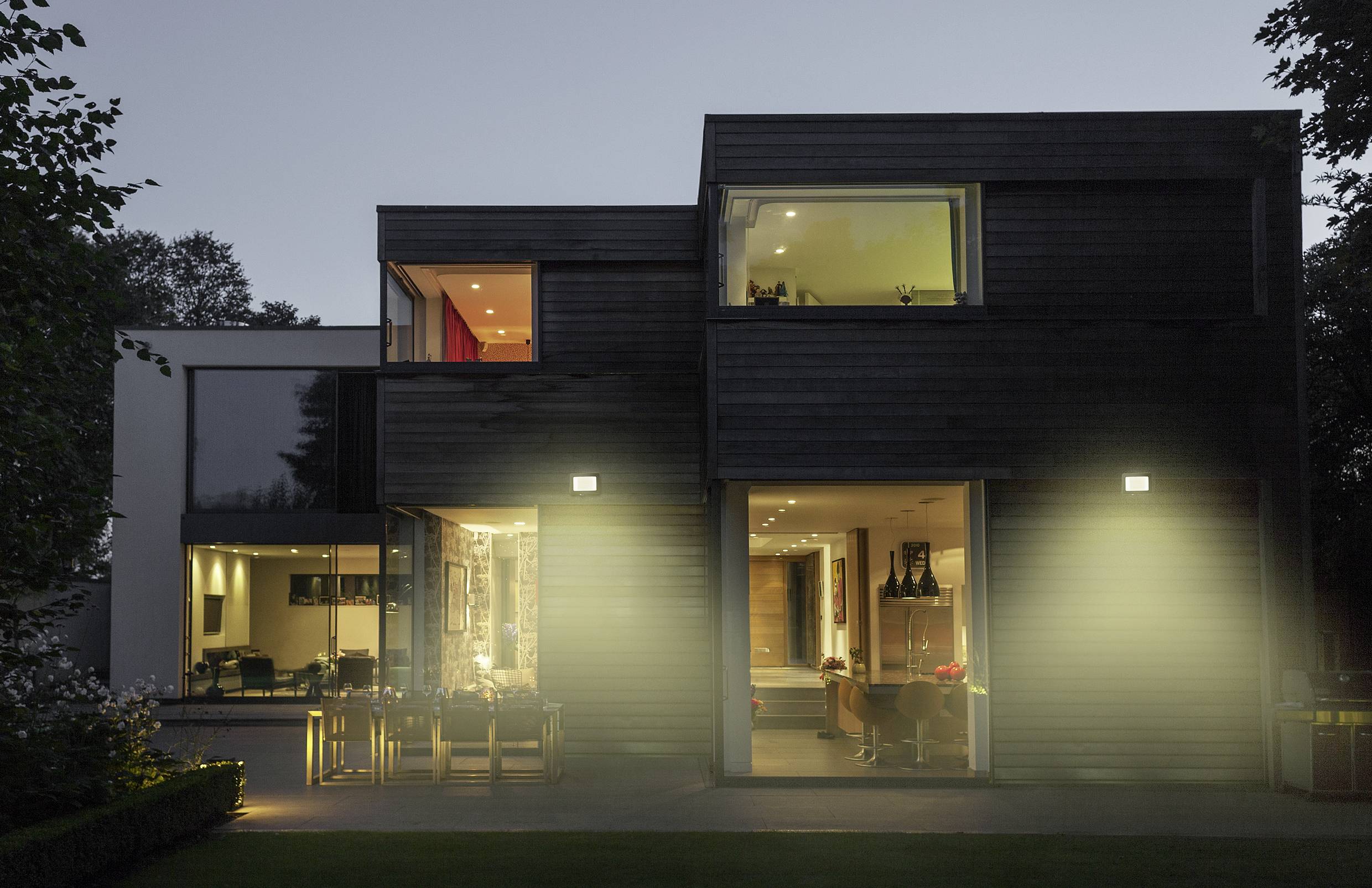 Une maison moderne au crépuscule, dotée de grandes fenêtres sur l'extérieur. La lumière émane de l'intérieur et illumine doucement le jardin.