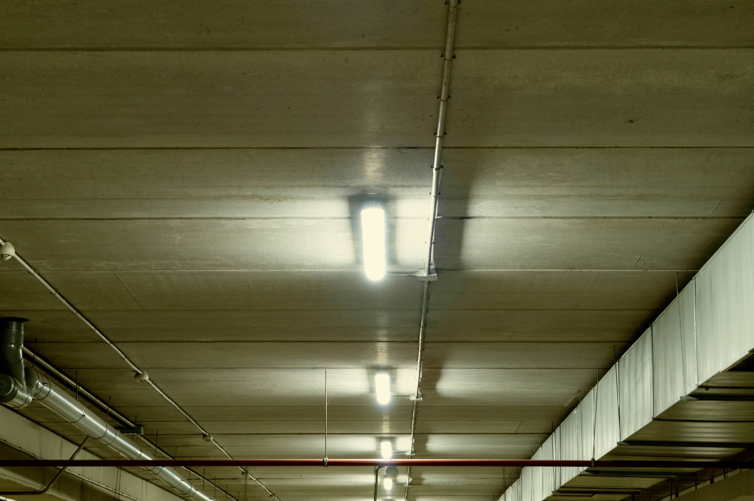 Vue du plafond d'un parking souterrain avec des dalles de béton et de longues tubes fluorescents. Des tuyaux courent parallèlement à l'éclairage.