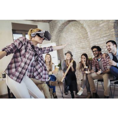 Une femme porte un casque de réalité virtuelle et bouge, tandis que cinq personnes rient en la regardant. Tous sont installés confortablement dans une pièce conviviale.