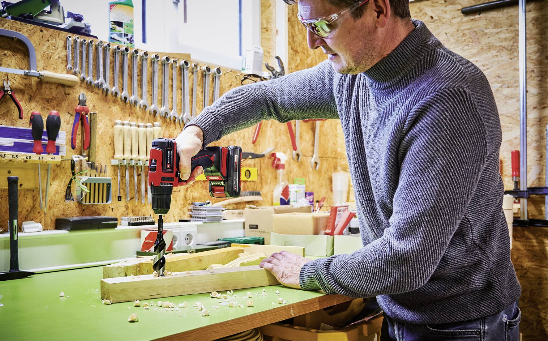 Un homme dans un atelier fore un morceau de bois avec une perceuse rouge, entouré d'outils accrochés au mur.