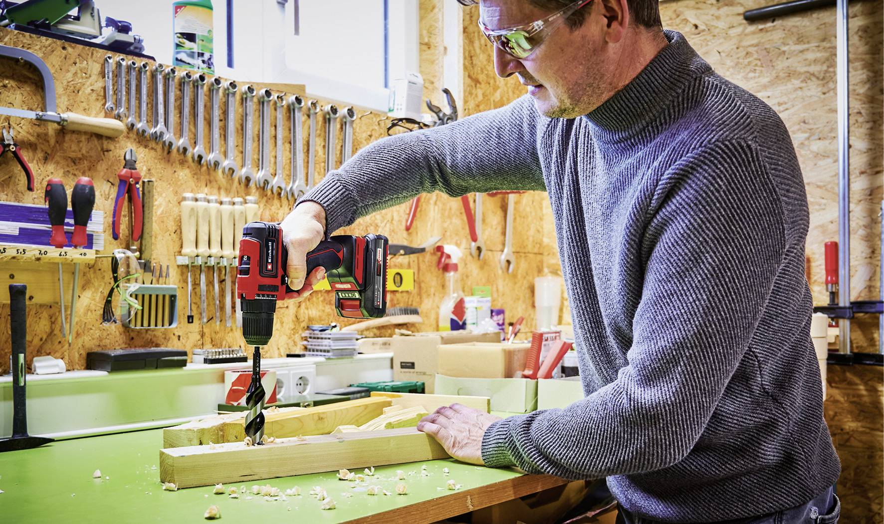 Un homme dans un atelier fore un morceau de bois avec une perceuse sans fil. En arrière-plan, des outils sont accrochés au mur, notamment des clés à molette et des marteaux.