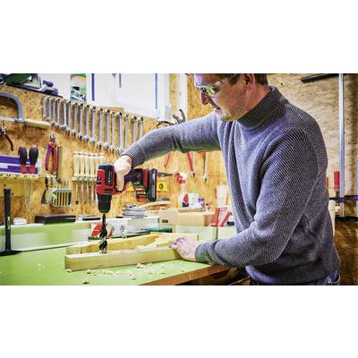 Un homme dans un atelier fore un morceau de bois avec une perceuse sans fil. En arrière-plan, des outils sont accrochés au mur, notamment des clés à molette et des marteaux.