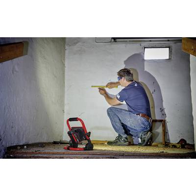 Un homme en t-shirt et jeans est agenouillé dans une pièce peinte en blanc et prend des mesures avec un mètre ruban. Une lampe de travail est posée au sol.