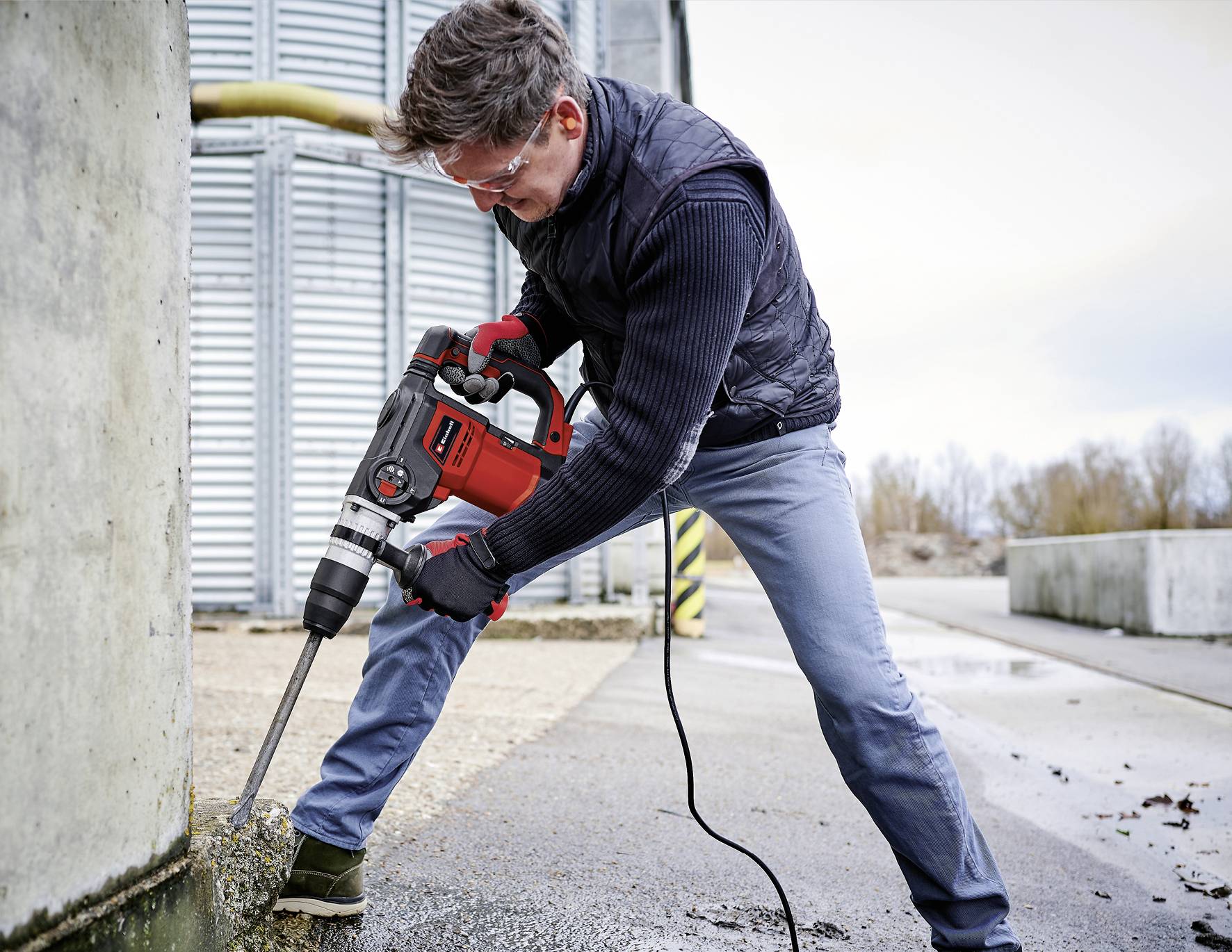 Une personne utilise un marteau-piqueur électrique sur du béton à l'extérieur. Elle porte des vêtements de protection. Des bâtiments industriels sont visibles en arrière-plan.