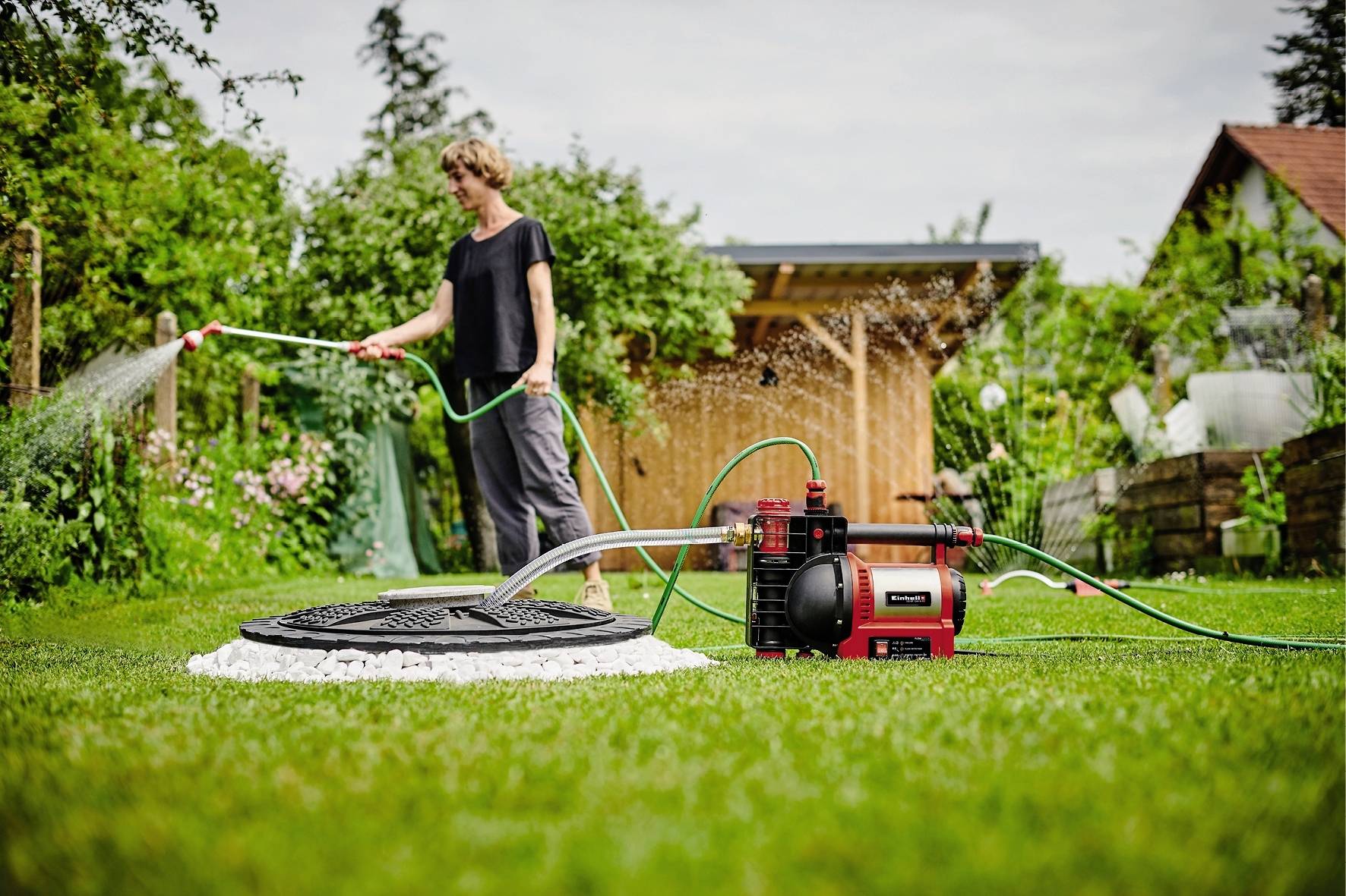 Une personne arrose la pelouse à l'aide d'un tuyau d'arrosage. Au premier plan, une pompe de jardin rouge est visible. À l'arrière-plan, on aperçoit des plantes.