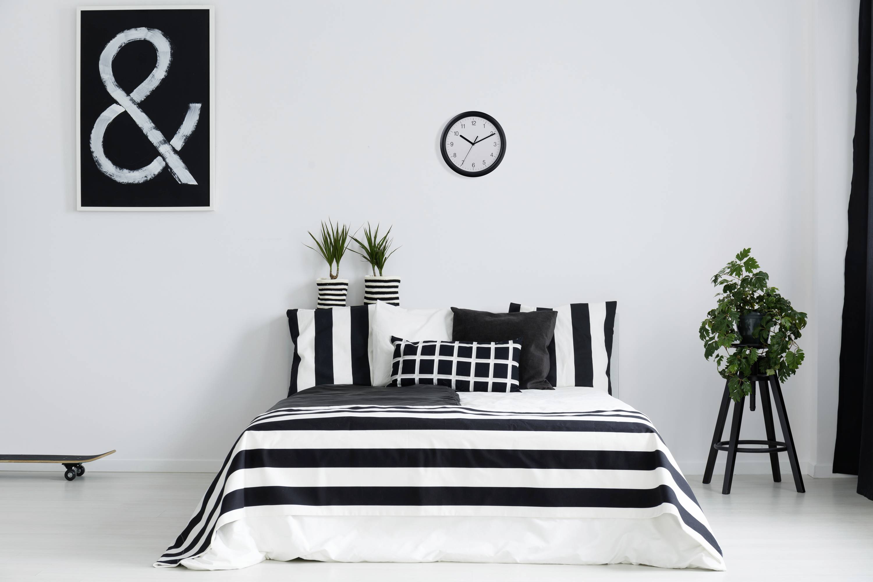 Une chambre moderne aménagée avec un schéma de couleurs blanc et noir, des draps rayés, deux plantes sur le côté de la tête de lit et une horloge murale.