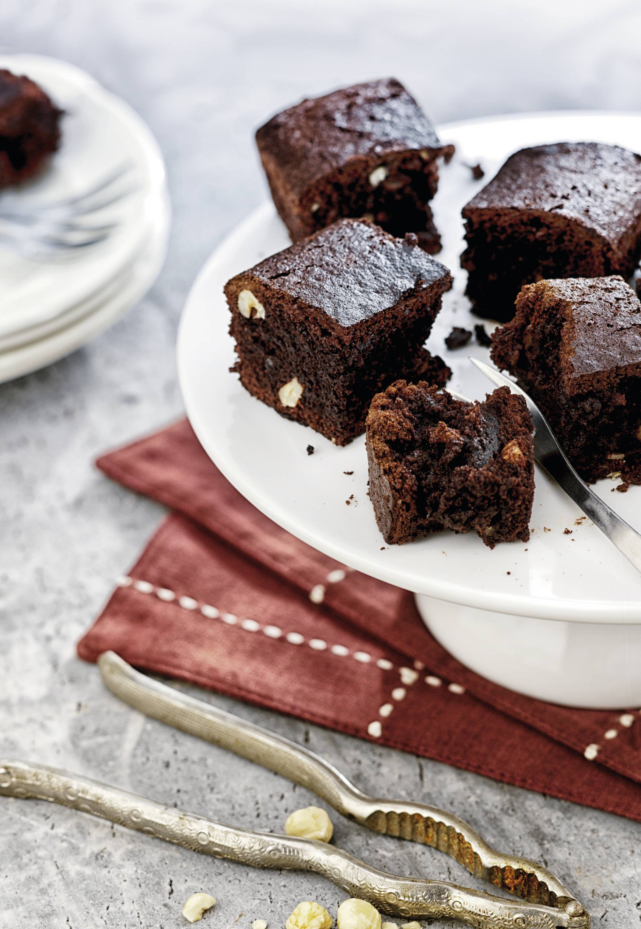 Une assiette avec plusieurs morceaux de gâteau au chocolat, garnis de noix, sur une serviette rayée rouge et blanc ; des assiettes en arrière-plan.