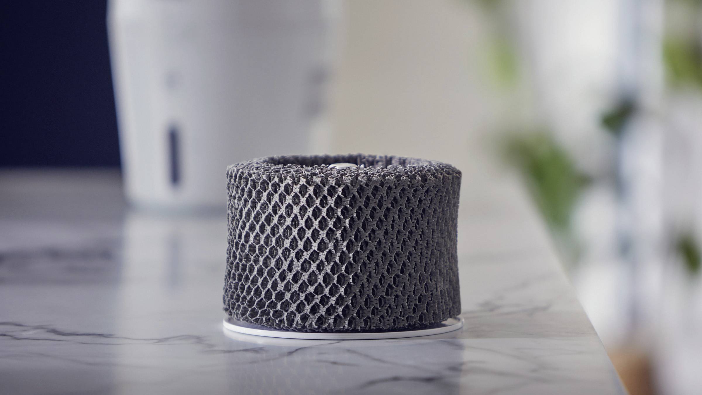 Filtre de purificateur d'air gris posé sur une table dans une pièce lumineuse. Le filtre est cylindrique et présente une structure en nid d'abeilles.