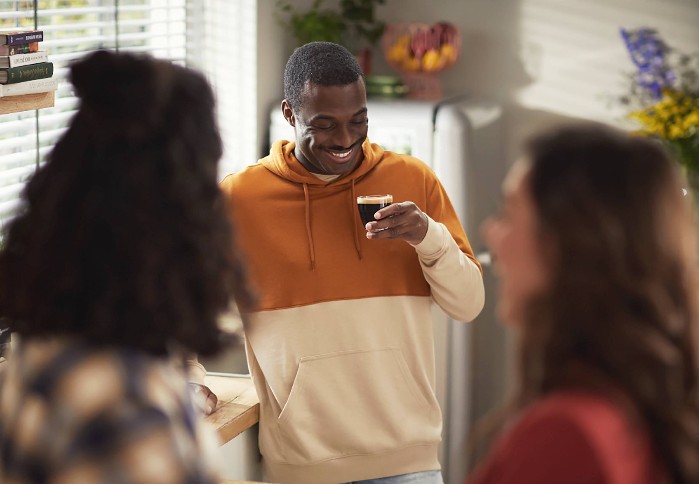 Un homme souriant portant un sweat à capuche orange tient une tasse de café dans la cuisine et parle à deux personnes à l'avant-plan.