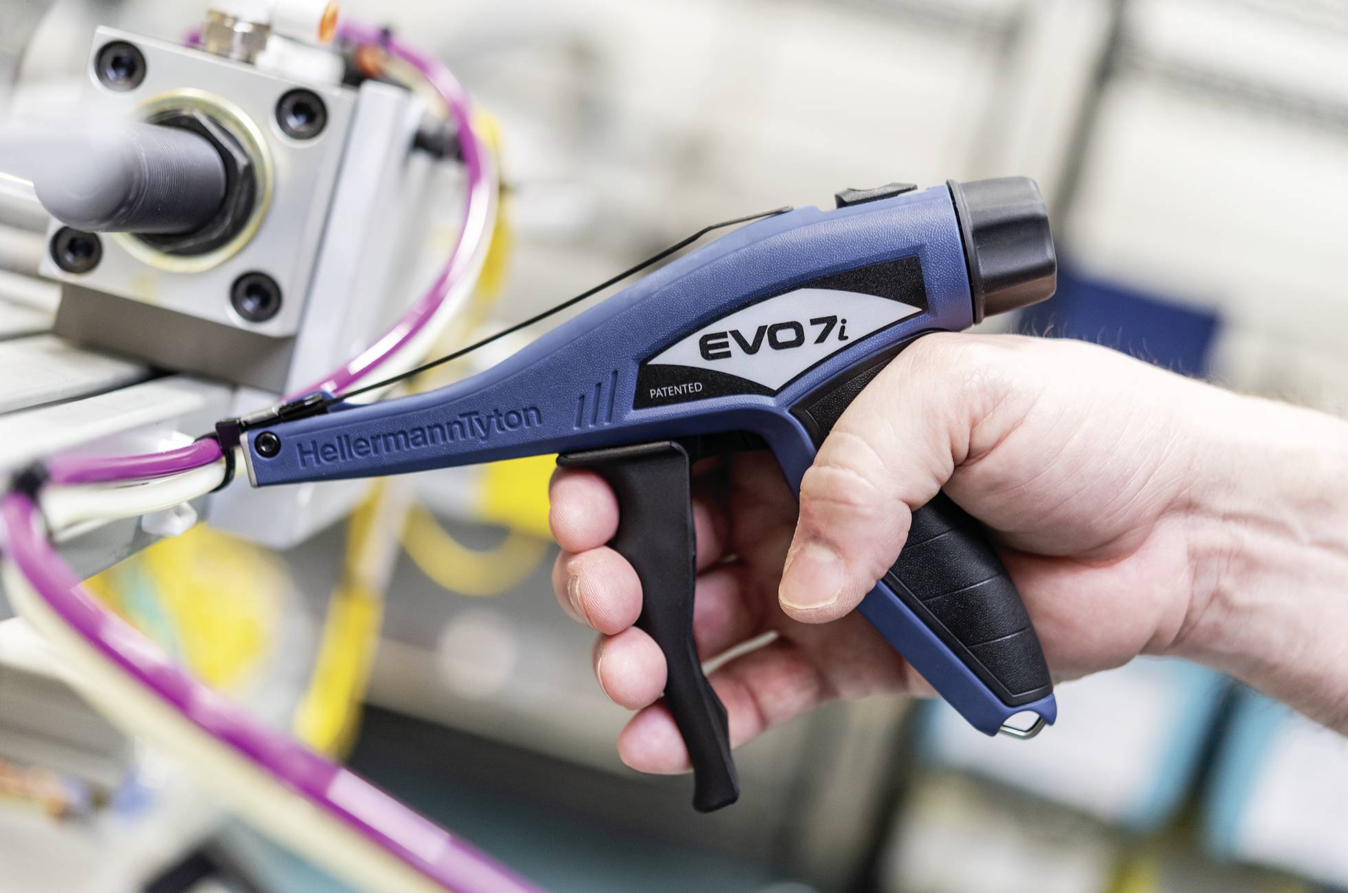 Une main tient un outil bleu en forme de pistolet portant l'inscription 'EVO 7i', alors qu'il travaille sur des câbles et un appareil.