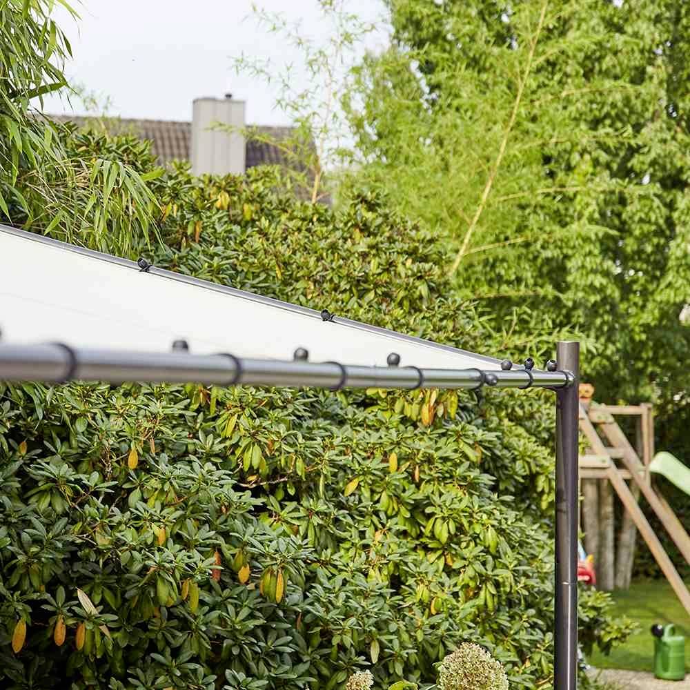 Un voile d'ombrage s'étend sur un jardin, entouré d'arbres et d'arbustes. Des toits de maisons sont visibles en arrière-plan.