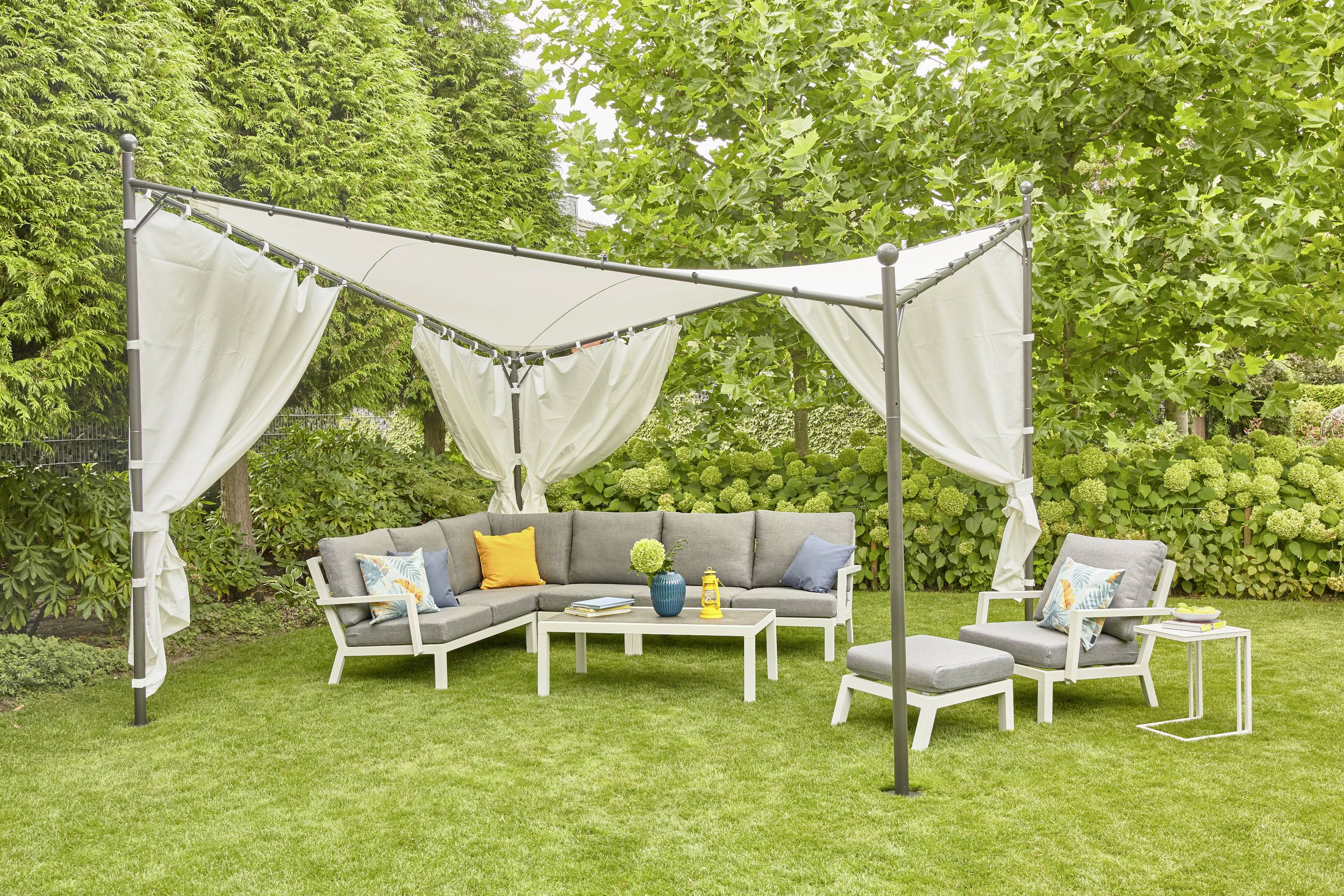 Un espace de détente de jardin avec des canapés gris, des coussins colorés et une voile d'ombrage. Entouré d'arbres et d'arbustes verts.