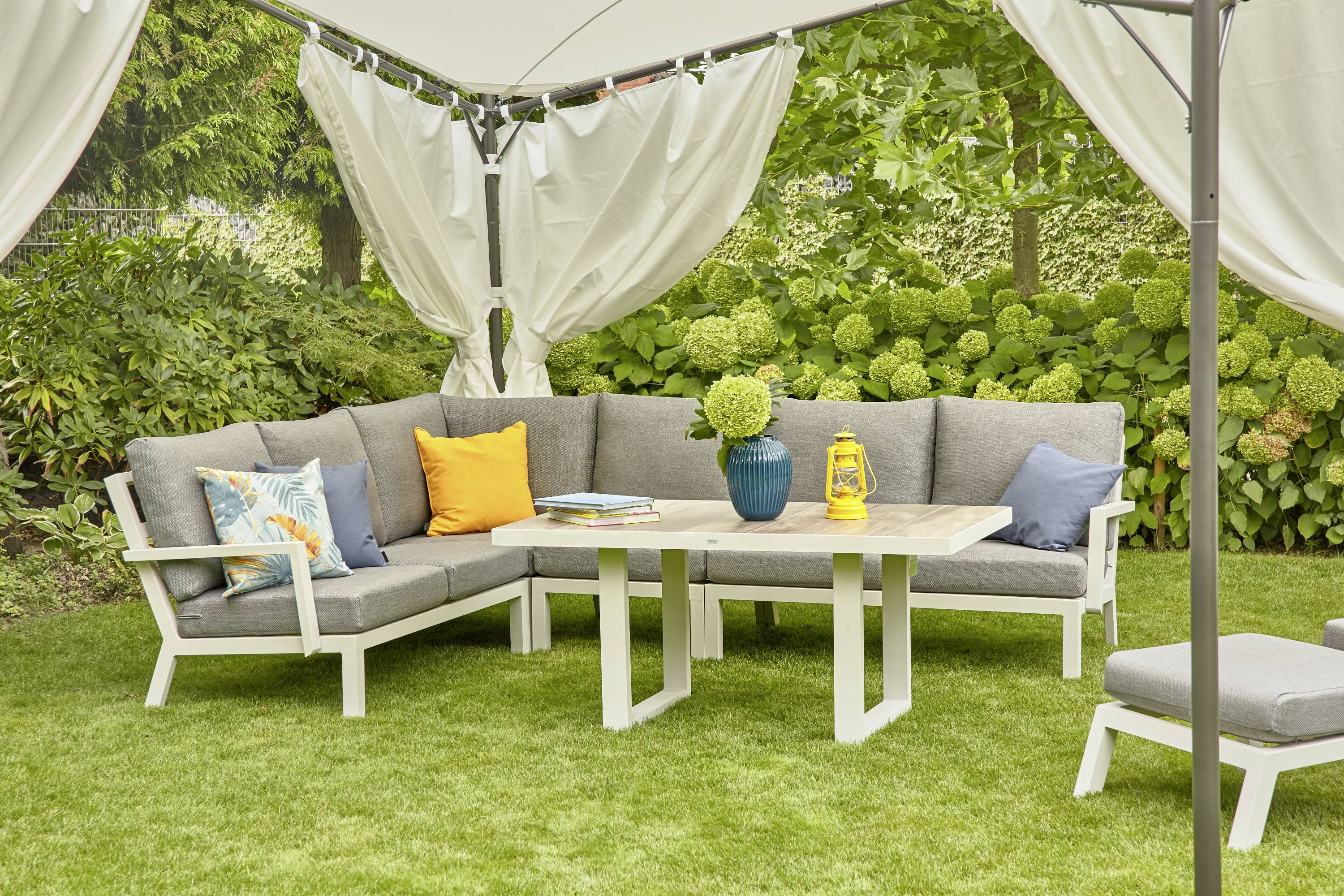 Jardin avec coin salon couvert : canapé d'angle gris avec coussins colorés, table décorée, végétation luxuriante en arrière-plan.