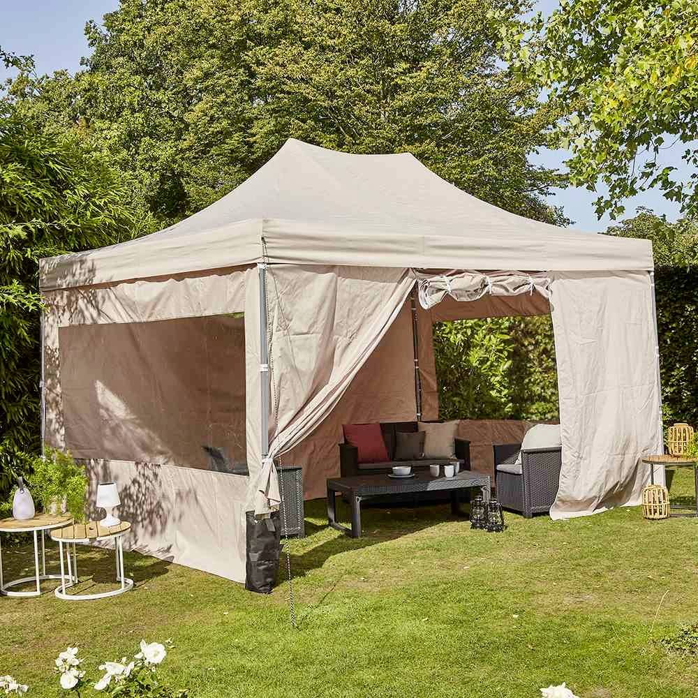 Un pavillon beige dans un jardin ensoleillé avec un coin salon, entouré de plantes et d'arbres. Idéal pour se détendre en plein air.
