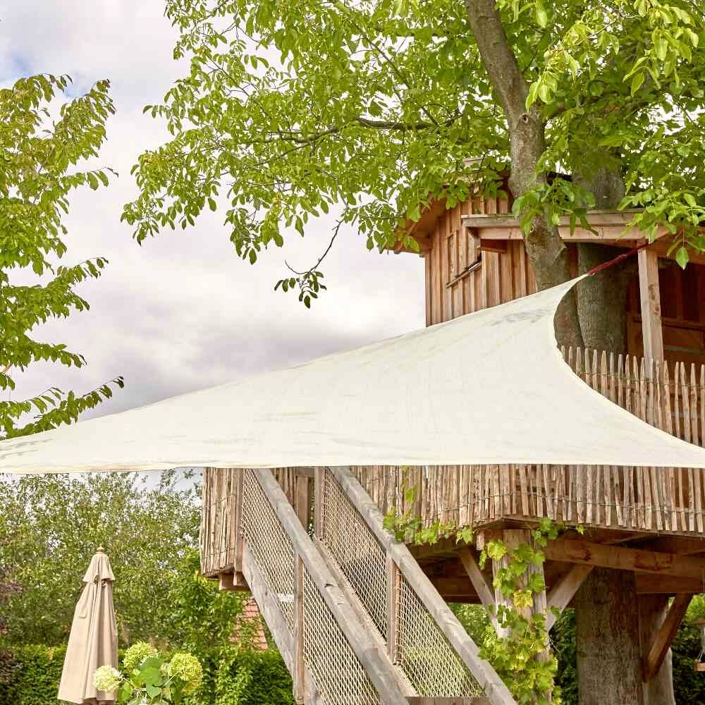 Cabane en bois perchée dans les arbres avec une échelle, entourée de feuilles vertes, révélant une ambiance de jardin ensoleillée.