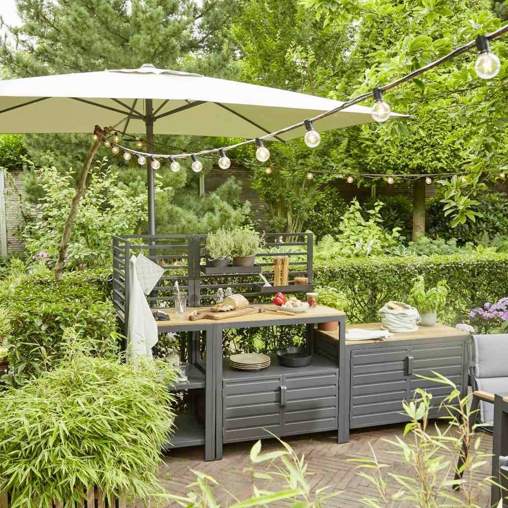 Un espace extérieur avec une zone de repas, un coin barbecue, des plantes et des guirlandes lumineuses. Un parasol offre de l'ombre au-dessus de la table.